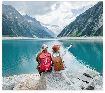 Two people sitting on rocky shore of lake and pointing to nearby mountains